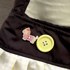 May include: Close-up of a brown fabric with a yellow gingham pattern and a yellow button. A small, pink and white dog-shaped embellishment is also visible. The fabric appears to be part of a garment or accessory.