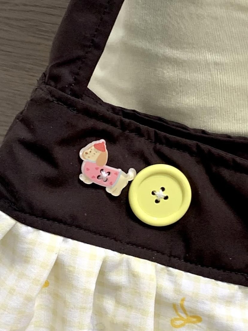 May include: Close-up of a brown fabric with a yellow gingham pattern and a yellow button. A small, pink and white dog-shaped embellishment is also visible. The fabric appears to be part of a garment or accessory.