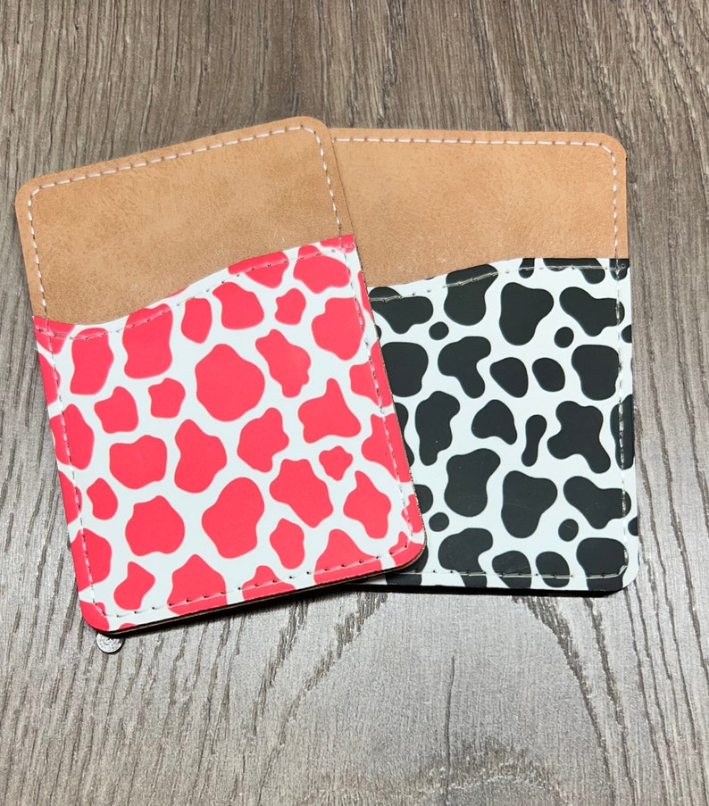 May include: Two phone card holders with a cow print design. One is red with white spots, and the other is black with white spots. Both have a tan pocket and white stitching.