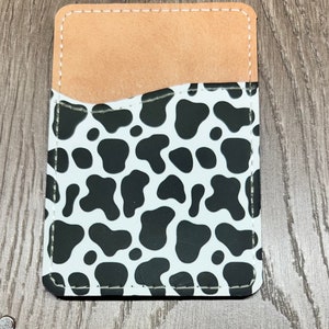 May include: A rectangular phone card holder with a cow print design. The holder features a white background with black spots, a tan pocket, and white stitching around the edges. The card holder is made of a durable material.