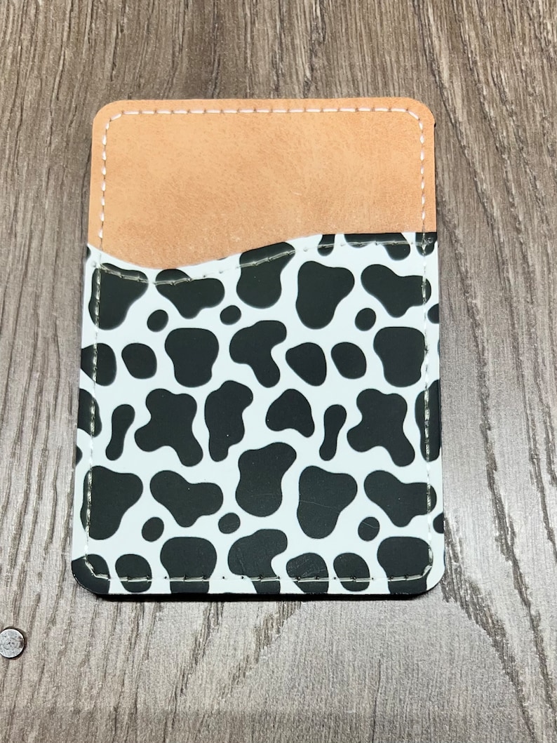 May include: A rectangular phone card holder with a cow print design. The holder features a white background with black spots, a tan pocket, and white stitching around the edges. The card holder is made of a durable material.