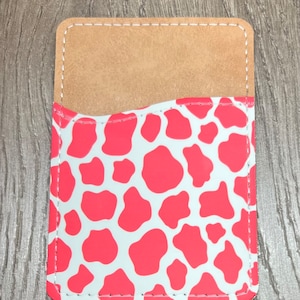 May include: A rectangular phone card holder with a light brown interior and a coral and white cow print exterior. The edges are stitched with white thread.