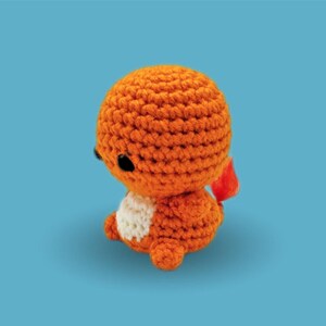 May include: A small, round, crocheted creature made of orange yarn with a cream-colored belly and two black bead eyes. The figure features a small, bright red yarn flame attached to its back, crafted with a textured, looped stitch pattern.