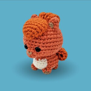 Könnte beinhalten: Ein kleines, handgefertigtes Häkel-Plüschtier in Fuchsform. Die Figur hat einen Körper aus leuchtend orangefarbenem Garn mit einem weißen Brustfleck, eine strukturierte orangefarbene Mähne, zwei spitze Ohren, einen geringelten Schwanz und zwei runde, glänzende schwarze Kunststoffaugen.