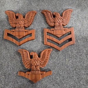 May include: Three wooden eagle insignia with chevron stripes. The eagle is carved in detail and the stripes are painted brown.