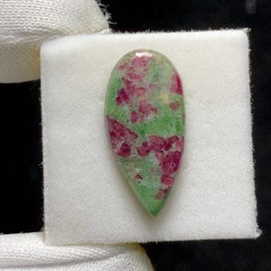 Puede incluir: Una gema en forma de lágrima con una base verde y vibrantes inclusiones rojas. La piedra pulida se presenta sobre un fondo blanco, mostrando sus variaciones de color y texturas naturales. Ideal para la creación de joyas.