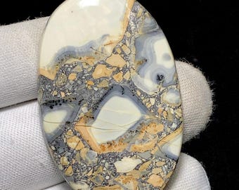 May include: Oval-shaped gemstone with a mosaic pattern. The stone features a blend of colors, including white, gray, tan, and black. The surface has a polished, glossy finish. The stone's design resembles a natural landscape or abstract art.