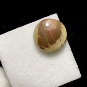 May include: A polished, round gemstone with a mix of brown and cream colors. The stone has a glossy surface and is set against a white fabric, with a black background. The gemstone's unique pattern adds to its visual appeal.