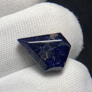 May include: A faceted, dark blue gemstone with a geometric shape. The stone has a polished, reflective surface with subtle variations in color and texture. The gemstone is set against a white background.