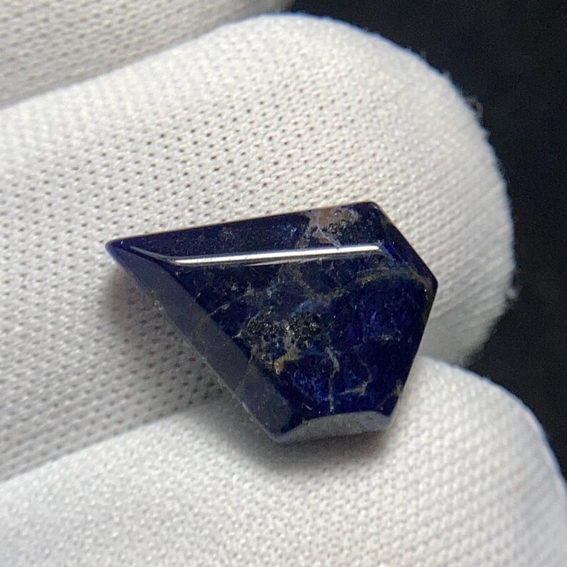 May include: A faceted, dark blue gemstone with a geometric shape. The stone has a polished, reflective surface with subtle variations in color and texture. The gemstone is set against a white background.