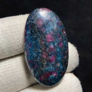 May include: An oval gemstone displaying a blend of blue and magenta colours. The stone has a speckled appearance, with magenta in small, irregular patches across a blue base. The surface is polished and glossy.