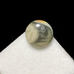 May include: A polished, round gemstone with a swirling pattern of cream, gray, and olive green. The stone has a glossy finish and a small area of orange-brown at the top. It sits on a white surface against a black background.