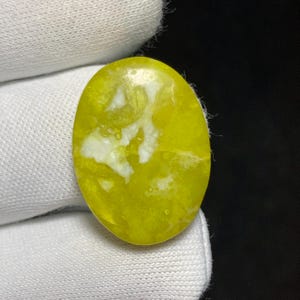 Peut inclure: Pierre précieuse ovale jaune vif, ornée d'un motif marbré blanc saisissant. La pierre est présentée sur un fond de tissu blanc, mettant en valeur sa surface polie et ses variations de couleurs uniques. La pierre est une pierre naturelle.