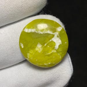 Pode incluir: Uma gema redonda e polida com um tom vibrante amarelo-esverdeado, acentuada por veios brancos. A pedra é segurada em uma mão enluvada branca, mostrando sua forma lisa e arredondada e seus padrões naturais. A superfície da gema reflete a luz.