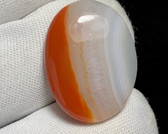 May include: Oval-shaped polished gemstone with bands of orange, white, and translucent sections. The stone is held against a white fabric background, showcasing its smooth, glossy surface. The gemstone's colors and patterns are clearly visible.