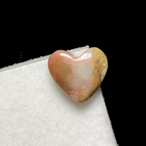 May include: A heart-shaped gemstone with a polished surface. The stone features a blend of colors, including shades of pink, brown, and cream. The heart is resting on a white surface against a black background. This item is suitable for jewelry making or as a decorative piece.
