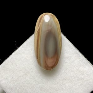 May include: An oval-shaped gemstone with a polished surface. The stone features a blend of colors, including beige, brown, and white, with a swirling pattern. The gemstone is displayed on a white cloth against a black background.