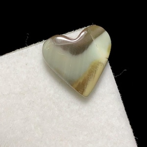 May include: A heart-shaped gemstone with a polished surface. The stone features a blend of colors, including shades of brown, cream, and a hint of pink. The heart is resting on a white surface, with a black background.