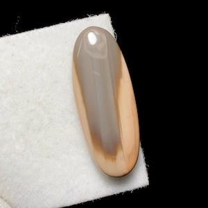 May include: An oval-shaped gemstone with a polished surface. The stone features a blend of colors, including light brown, gray, and tan, creating a striped pattern. The gemstone is set against a white background and a black backdrop.