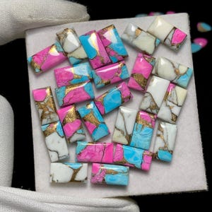Puede incluir: Colección de cabujones de piedras preciosas rectangulares en varios colores. Las piedras presentan una mezcla de rosa, turquesa, blanco y dorado. Las gemas están dispuestas sobre una superficie blanca, mostrando sus vibrantes colores.
