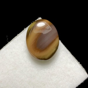 May include: Oval-shaped gemstone with a polished surface. The stone features a blend of brown, tan, and yellow hues, creating a striped pattern. The gemstone is set against a white surface with a black background.