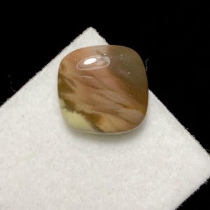 May include: A polished, square-shaped gemstone with a mix of brown, tan, and cream colors. The stone has a smooth, glossy surface and is set on a white, textured surface against a black background. The gemstone's unique color patterns are visible.