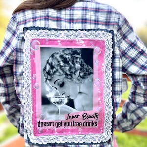 May include: A white and blue plaid flannel shirt with a pink and white lace trim. The back of the shirt features a pink patch with a black and white photo of a woman drinking from a glass. The text "Inner Beauty doesn't get you free drinks" is printed on the patch.