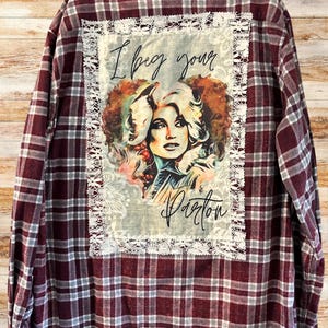 May include: A red and white plaid flannel shirt with a white lace trim around a patch featuring a portrait of Dolly Parton with the text "I beg your Parton".