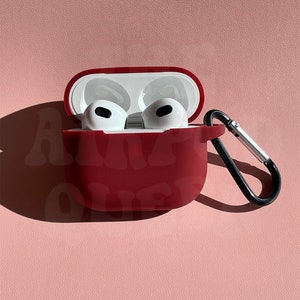 Pode incluir: Uma capa de silicone vermelha para fones de ouvido sem fio com um mosquetão. A capa está aberta e os fones de ouvido estão dentro.