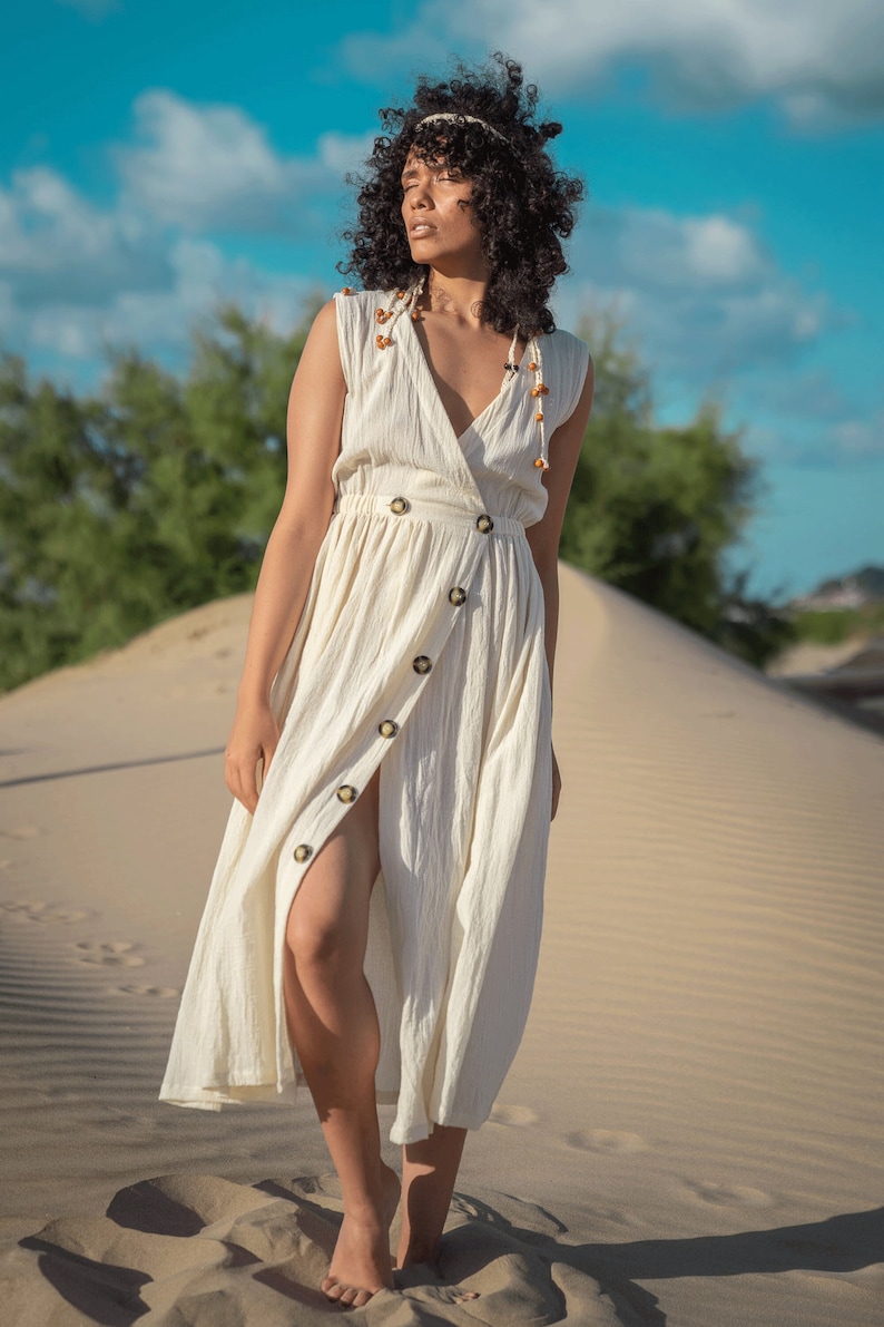 K&ouml;nnte beinhalten: Eine Frau in einem wei&szlig;en Leinenkleid mit Knopfleiste und einer Perlenkette. Sie steht in einer W&uuml;stenlandschaft mit Sandd&uuml;nen im Hintergrund.
