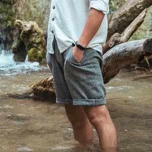 May include: A man wearing a white linen shirt and gray linen shorts stands in a shallow stream. He is wearing a black fitness tracker on his wrist.