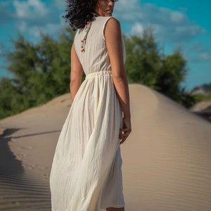 K&ouml;nnte beinhalten: Eine Frau in einem wei&szlig;en Maxikleid mit einer Perlenkette und einem Stirnband geht an einem Sandstrand entlang.