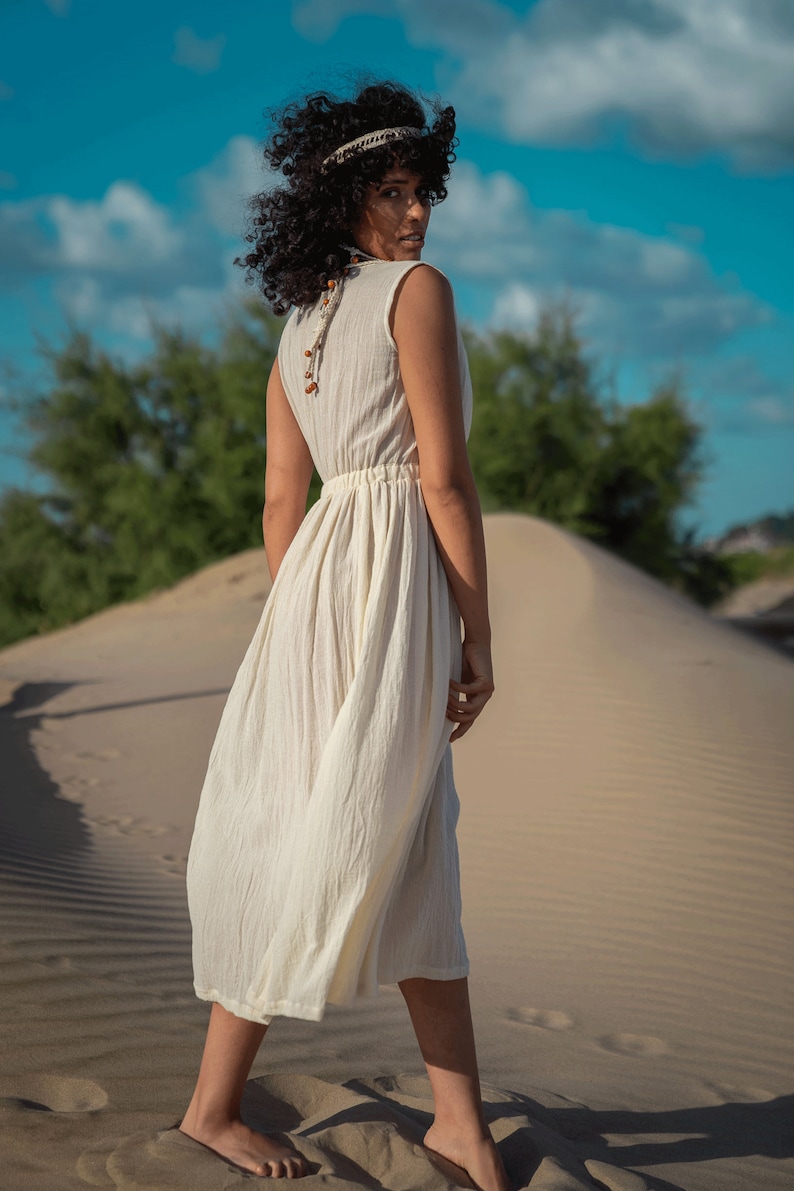 K&ouml;nnte beinhalten: Eine Frau in einem wei&szlig;en Maxikleid mit einer Perlenkette und einem Stirnband geht an einem Sandstrand entlang.