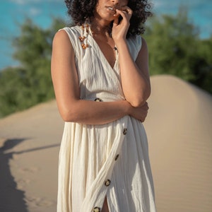 K&ouml;nnte beinhalten: Eine Frau mit lockigem, dunklem Haar tr&auml;gt ein wei&szlig;es, &auml;rmelloses Kleid mit einem Schlitz vorne und Kn&ouml;pfen. Sie steht in einer W&uuml;stenlandschaft mit Sandd&uuml;nen.