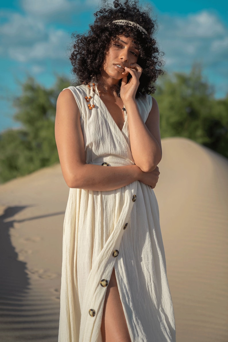 K&ouml;nnte beinhalten: Eine Frau mit lockigem, dunklem Haar tr&auml;gt ein wei&szlig;es, &auml;rmelloses Kleid mit einem Schlitz vorne und Kn&ouml;pfen. Sie steht in einer W&uuml;stenlandschaft mit Sandd&uuml;nen.