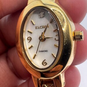 May include: A gold-tone oval-shaped watch with a white face and black hands. The watch has a diamond at the 12 o'clock position and the words "ELGIN DIAMOND" on the face.