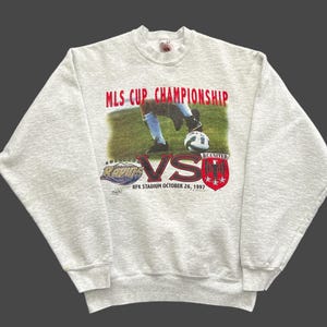 May include: A light gray sweatshirt featuring a soccer match graphic. The graphic includes the text "MLS CUP CHAMPIONSHIP" in red, a soccer player kicking a ball, and the words "RAPIDS" and "UNITED" with a "VS" in the middle. The date "OCTOBER 26, 1997" is also visible.