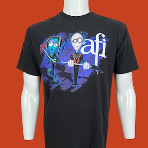 May include: A black short-sleeve t-shirt featuring a graphic print of two stylized cartoon characters. One character holds a red umbrella, and the other holds a long tool. The text "afi" is printed in white in the upper right corner of the graphic.
