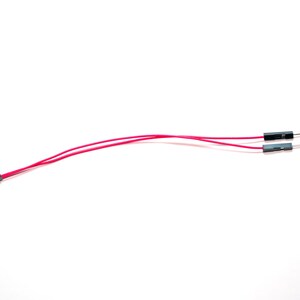 May include: A jumper wire assembly featuring two thin red insulated electrical wires. One end of the assembly has a single black female connector, while the other end splits into two separate black male pin connectors for electronic circuit connections.