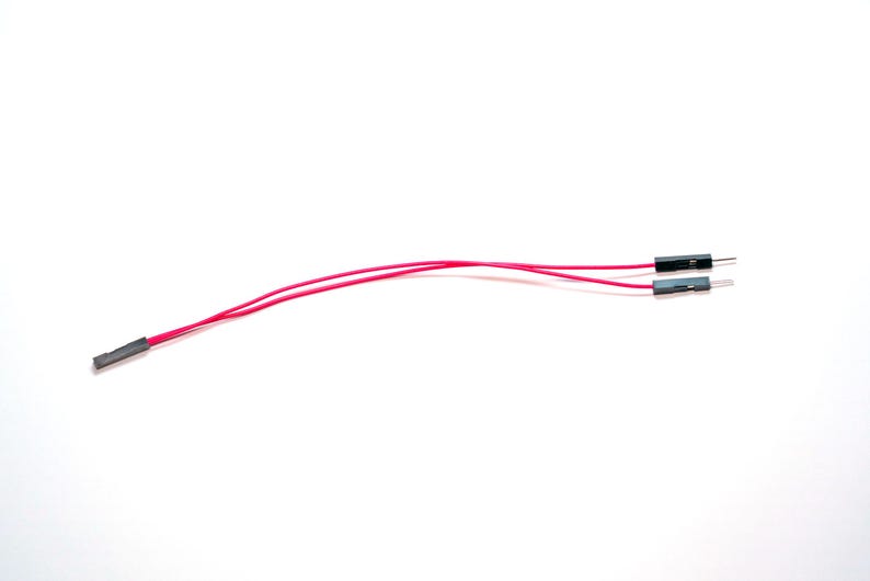 May include: A jumper wire assembly featuring two thin red insulated electrical wires. One end of the assembly has a single black female connector, while the other end splits into two separate black male pin connectors for electronic circuit connections.