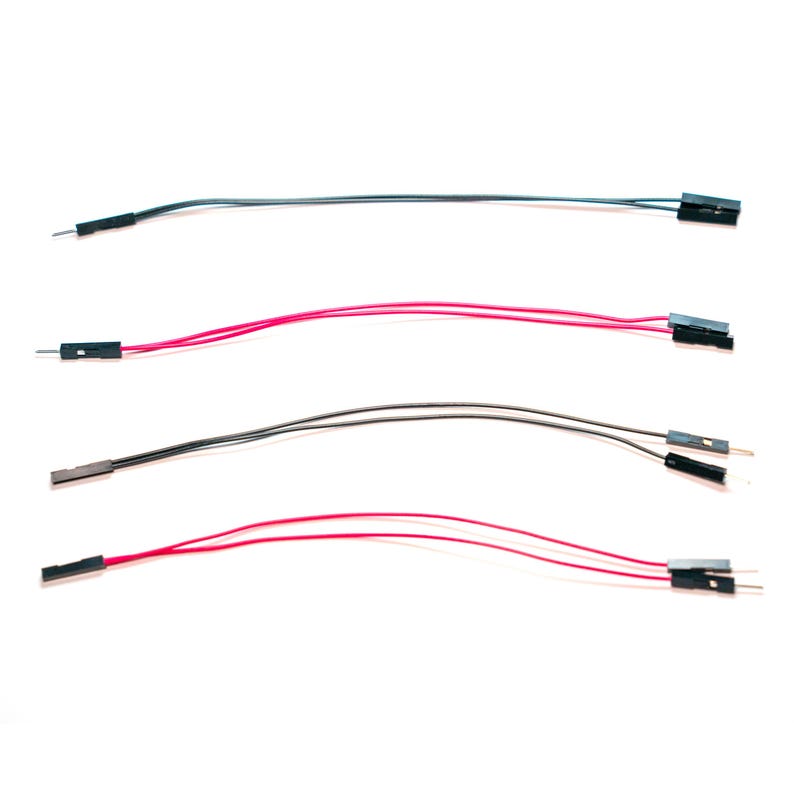 May include: Four jumper wires arranged horizontally. Each wire features a flexible insulated cord in either black or red, with a black plastic connector housing at both ends. The connectors contain exposed metal pins or sockets for electronic connections.