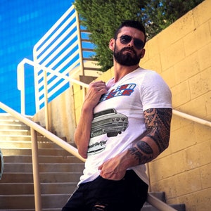May include: A white t-shirt with a vintage car graphic and the text "70's Ride" printed on the front. The man is wearing black jeans and sunglasses.