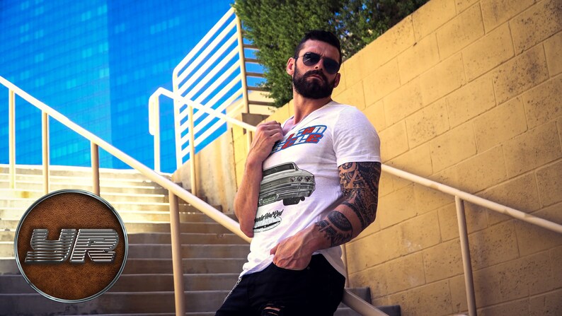 May include: A white t-shirt with a vintage car graphic and the text "70's Ride" printed on the front. The man is wearing black jeans and sunglasses.