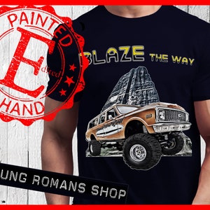 Könnte beinhalten: Ein marineblaues T-Shirt mit einer braunen und gelben Illustration eines alten Geländewagens, der einen Berg hinauf fährt. Der Text "Blaze the Way" ist in Gelb über dem Fahrzeug gedruckt. Das T-Shirt hat einen abgenutzten roten Stempel, auf dem "Painted by Hand" in weißen Buchstaben steht.