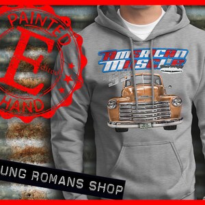 Peut inclure: Un sweat à capuche gris avec un motif de camion vintage et le texte "American Muscle" en bleu et rouge. Le camion est brun et a une plaque d'immatriculation blanche qui dit "Made in USA". Le sweat-shirt a une poche sur le devant.