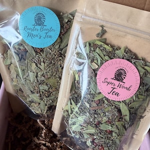 May include: Two clear plastic bags filled with dried herbs and leaves. Each bag has a round sticker with text. One says "Rooster Booster Men's Tea" in blue, the other says "Super Womb Tea" in pink. The bags are in a brown box.