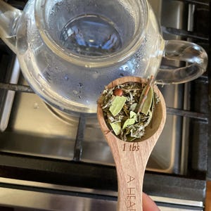 Puede incluir: Una cuchara de madera con una cucharada colmada de hierbas secas, incluyendo hojas verdes y pequeñas bayas marrones. La cuchara está etiquetada como "A Heap 1 tbs".