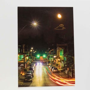 May include: A nighttime street scene with cars and buildings illuminated by streetlights. The long exposure captures streaks of red and orange light from moving vehicles, creating a dynamic visual effect. The sky is dark with a bright moon.