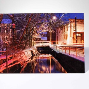 May include: A rectangular print of a winter scene with a snow-covered river reflecting the lights of buildings and street lamps. The sky is a deep blue, and the overall color palette is a mix of cool and warm tones.