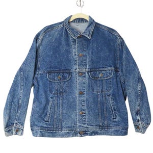 May include: A blue acid-wash denim jacket featuring a pointed collar, long sleeves with buttoned cuffs, and a front button closure. The jacket includes two chest pockets with buttoned flaps and two vertical side pockets, all in a classic denim fabric.
